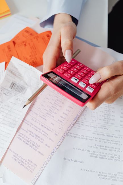 Close-up of a person's hands holding a pink calculator while reviewing paperwork, including receipts, invoices, and packing lists, on a white surface. An orange printed document and a pencil are also visible among the papers. The scene appears to be inside a property or office space, with the focus on calculations related to a home relocation or moving logistics. This image illustrates the process of estimating costs and managing packing and moving expenses, relevant to house removals services offered by Movers Bermondsey, as seen on their website's Bermondsey removals cost calculator page.
