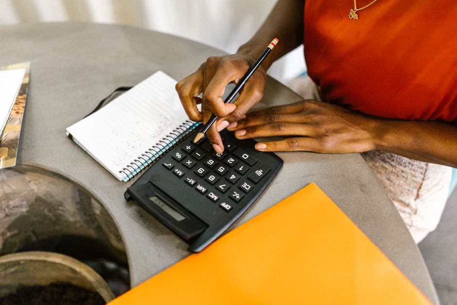 Close-up of a person with dark skin using a black calculator with white buttons, accompanied by a pen held in their right hand and a notepad with handwritten notes on a grey surface. The individual is wearing a red shirt and has their left hand resting on the calculator. Nearby, there is an orange folder or document, and a large potted plant pot is partially visible in the foreground. This setting suggests a planning or calculation scene related to house removals or moving logistics, with the focus on financial or cost assessment for a home relocation. The environment appears to be indoors with neutral lighting, and the scene is relevant to furniture transport and packing and moving activities often managed by companies like Movers Bermondsey, as referenced on the Bermondsey removals cost calculator page.