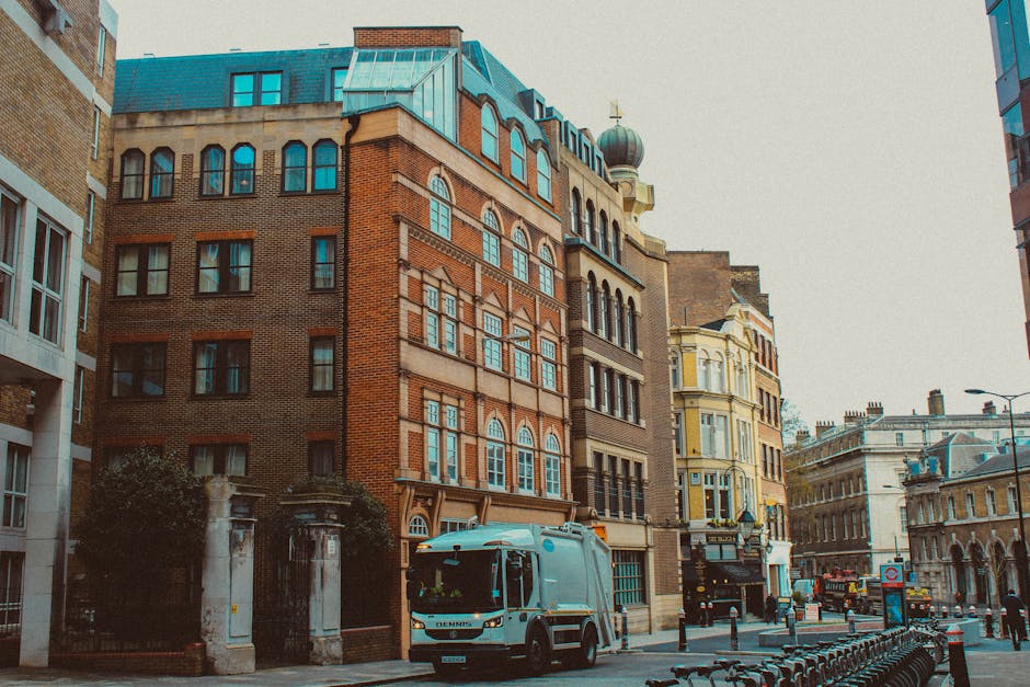 A street scene showing a delivery van parked on the sidewalk in front of a row of multi-storey residential and commercial buildings with historic architectural details, including large windows and orn